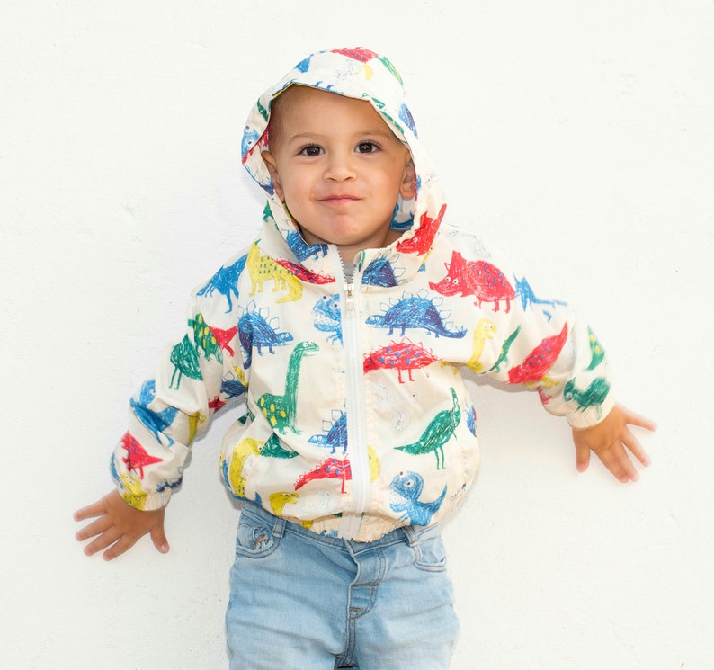 May include: A white hooded jacket with a colorful dinosaur print. The jacket has a zipper closure and is made of a lightweight fabric.