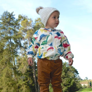 May include: A young child wearing a white jacket with a dinosaur print, brown corduroy pants, and black sneakers. The child is wearing a white knit hat with a pom-pom on top. The child is standing on a green grassy field.