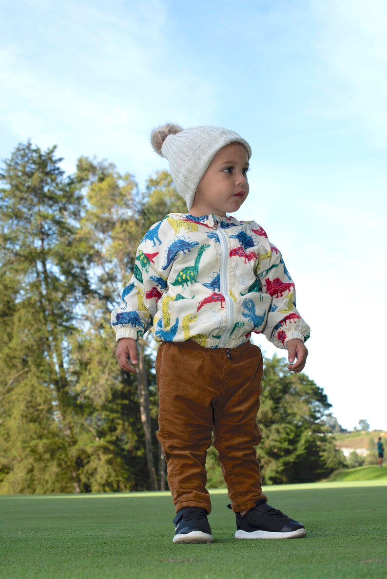 May include: A young child wearing a white jacket with a dinosaur print, brown corduroy pants, and black sneakers. The child is wearing a white knit hat with a pom-pom on top. The child is standing on a green grassy field.