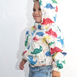 May include: A young child wearing a white jacket with a colorful dinosaur print. The jacket has a hood and a zipper closure. The child is wearing blue jeans.
