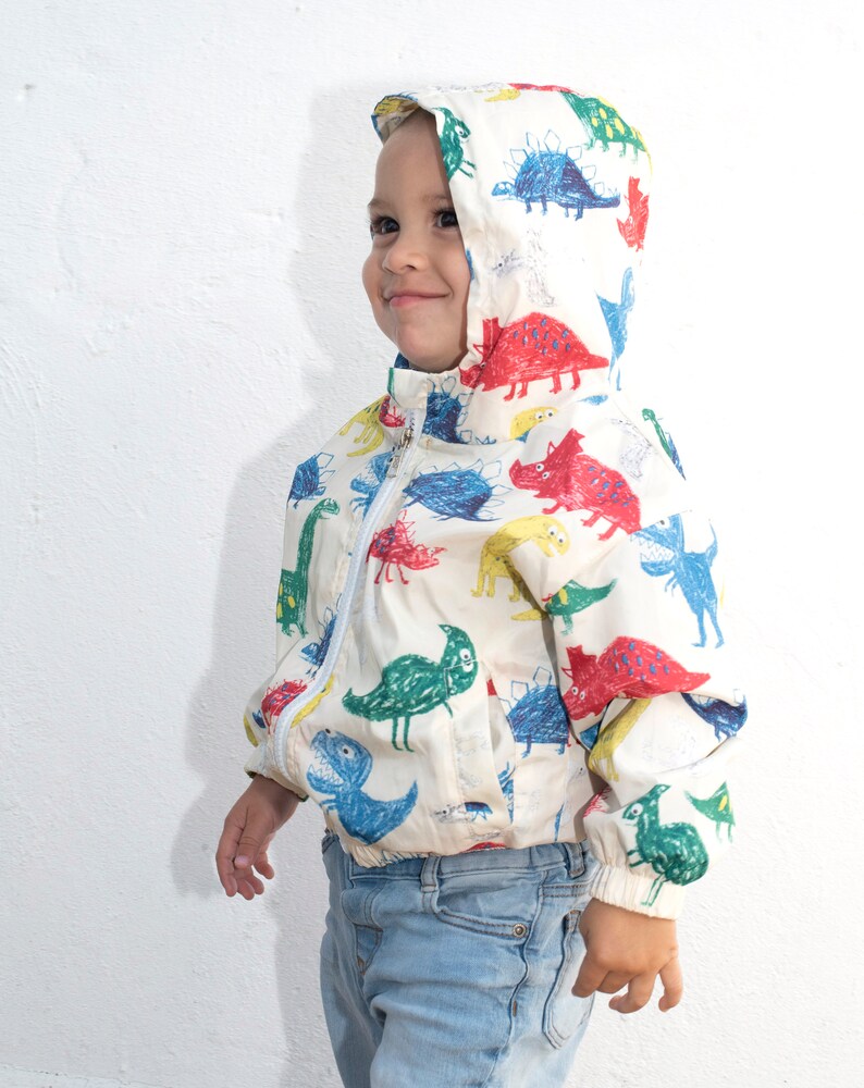 May include: A young child wearing a white jacket with a colorful dinosaur print. The jacket has a hood and a zipper closure. The child is wearing blue jeans.