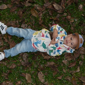 May include: A young child wearing a white jacket with a dinosaur print, blue jeans, and white sneakers lies on a bed of green grass and brown leaves.