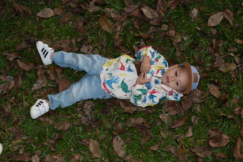 May include: A young child wearing a white jacket with a dinosaur print, blue jeans, and white sneakers lies on a bed of green grass and brown leaves.