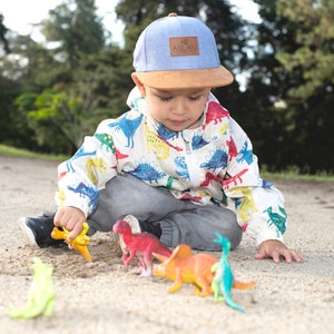 May include: A young child wearing a blue and brown baseball cap with the word "ASLAN" on it, sits on the ground playing with colorful dinosaur toys.