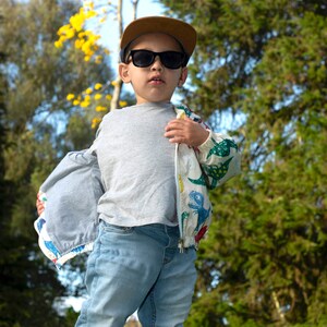 May include: A young person wearing a grey t-shirt, blue jeans, a brown baseball cap, and a white jacket with a colorful dinosaur print. The jacket is unzipped and the person is looking at the camera.
