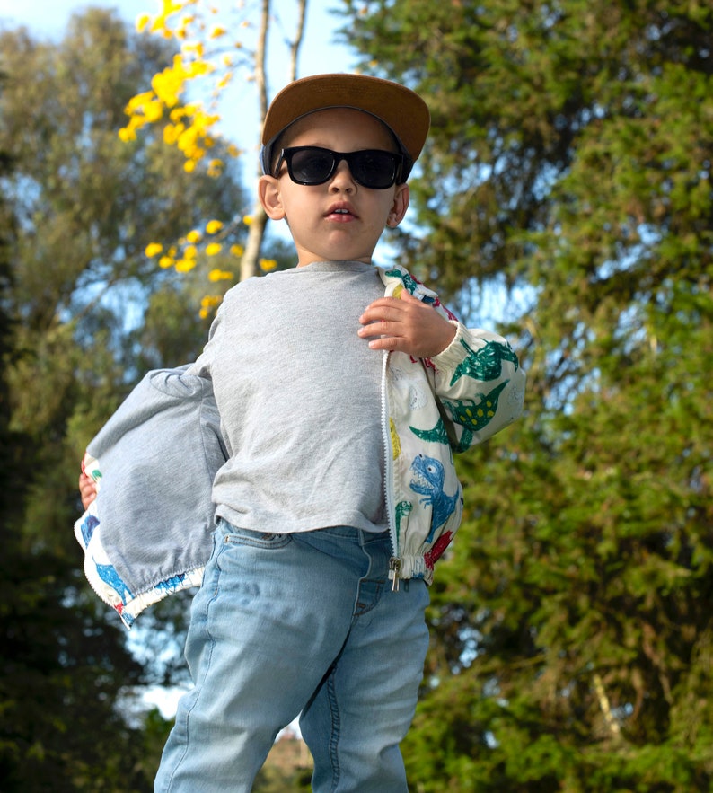 May include: A young person wearing a grey t-shirt, blue jeans, a brown baseball cap, and a white jacket with a colorful dinosaur print. The jacket is unzipped and the person is looking at the camera.