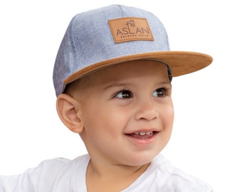 May include: A young child wearing a gray and brown baseball cap with the text "ASLAN BELOVED CHILD" on the front.