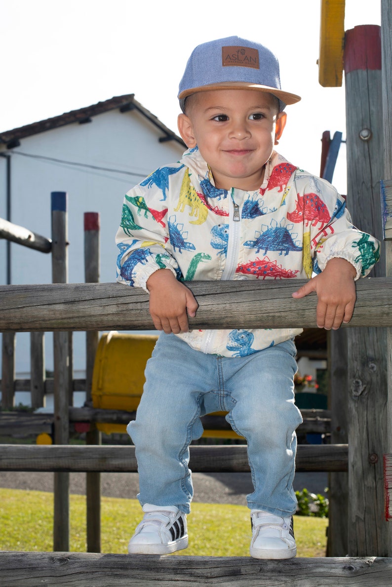 May include: A young child wearing a blue and brown baseball cap with the word "ASLAN" on it. The child is wearing a white jacket with a colorful dinosaur print and blue jeans. The child is standing on a wooden platform.