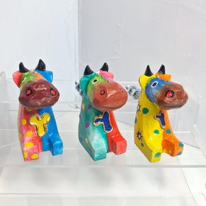 May include: Three brightly painted wooden cow ornaments. Each cow features a different colour palette, including red, blue, yellow, and green, with gold detailing. The cows have black horns and are displayed on a clear shelf.