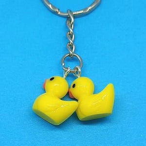 May include: A yellow duck keyring featuring two small, glossy yellow duck figurines. Each duck has a black eye and an orange beak. The ducks are attached to a silver-coloured metal chain and ring, set against a solid blue background.