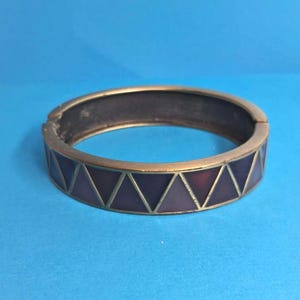 May include: A hinged bangle bracelet featuring a metallic gold-tone finish and a repeating geometric pattern of dark purple and maroon triangles. The bracelet has a smooth, rounded interior and a secure clasp mechanism visible on the side.