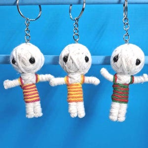May include: Three handmade keyrings featuring small, white yarn dolls with black button eyes and outstretched arms. Each doll wears a striped top in red and yellow, orange and yellow, or green and red. The keyrings hang against a blue background.