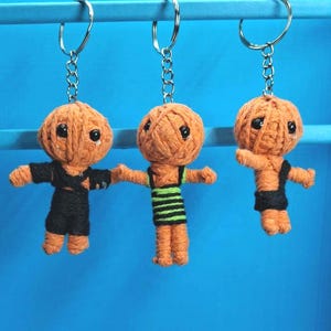 May include: Three handmade keyrings featuring orange yarn doll figures with black button eyes and black clothing. The dolls are hanging from silver key rings against a blue background. One doll has a green and black striped shirt.