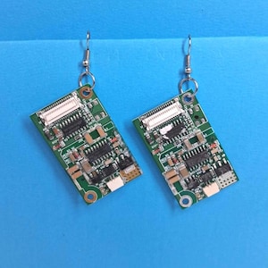 May include: A pair of quirky earrings crafted from recycled circuit boards. Each earring displays a rectangular green board with diverse electronic components, such as chips and connectors. The earrings are suspended from silver-coloured fish hook ear wires.