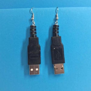 May include: A pair of quirky earrings crafted from recycled black USB cables. Each earring has a silver hook and a black USB connector, presenting a creative, tech-themed design. The earrings are set against a blue backdrop.