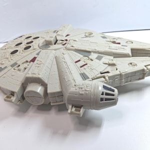 May include: A detailed, off-white toy model of the Millennium Falcon starship. The model features intricate paneling, grey and red accents, and a distinctive circular cockpit. This collectible item is a replica from the Star Wars franchise.