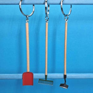 May include: Three miniature garden tool keyrings suspended from a light blue bar. The tools are a red shovel, a green rake, and a black hoe, each with a wooden handle and a silver keyring.