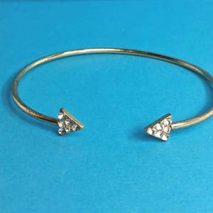 May include: A gold-toned open cuff bracelet featuring a thin, circular band. Each end of the bracelet terminates in a small, flat triangle shape. These triangular ends are embellished with multiple clear, sparkling rhinestones.