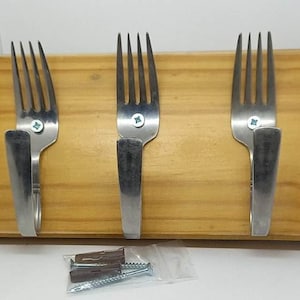 May include: A wooden wall-mounted rack with three silver forks mounted as hooks. The rack is made of light wood and has a natural finish. The forks are attached to the wood with screws.