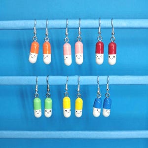 May include: A selection of novelty earrings shaped like smiling pills, in various colours. Each earring hangs from a silver hook. The earrings are displayed against a blue backdrop, with colours including orange, pink, red, green, yellow, and blue.