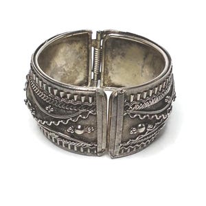 May include: Silver hinged cuff bracelet with detailed embossed patterns. The bracelet features intricate designs including triangles, chain links, and small beaded accents. The clasp is visible at the top, set against a white background.