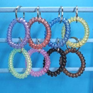 May include: A selection of spiral hair ties in a range of colours: purple, pink, blue, yellow, green, black, and brown. Each tie is attached to a silver metal ring. The background is a solid light blue.