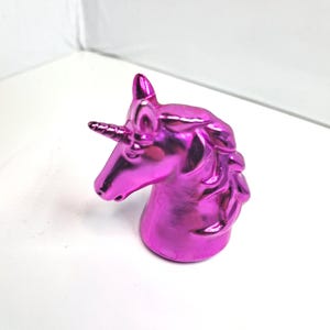 May include: A metallic magenta unicorn head figurine. The sculpture features a detailed mane, a pointed horn, and a smooth, reflective surface. The unicorn is set against a white background.