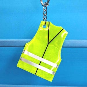 May include: A neon yellow vest-shaped keyring with two silver reflective stripes and black lines. The keyring is attached to a silver chain and ring. The background is a solid blue.
