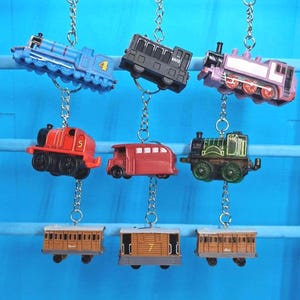 May include: A selection of toy train keyrings in various colours, including blue, red, green, and purple. Each keyring features a different train design, with some displaying numbers. The keyrings are suspended from silver chains.