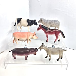 May include: Six plastic farm animal figurines: a black and white cow, a grey sheep, a pink pig, a maroon horse, a purple hippo, and a tan goat. The animals are arranged on clear, tiered shelves against a white backdrop.