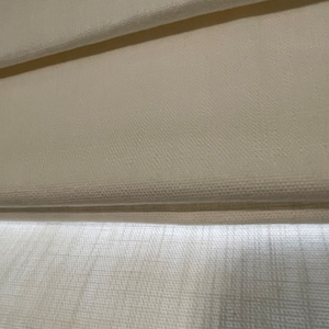Puede incluir: Primer plano de una cortina romana de color crema, con una tela texturizada con pliegues horizontales. La cortina est&aacute; parcialmente bajada, revelando la construcci&oacute;n en capas y el sutil juego de luces y sombras en la superficie.