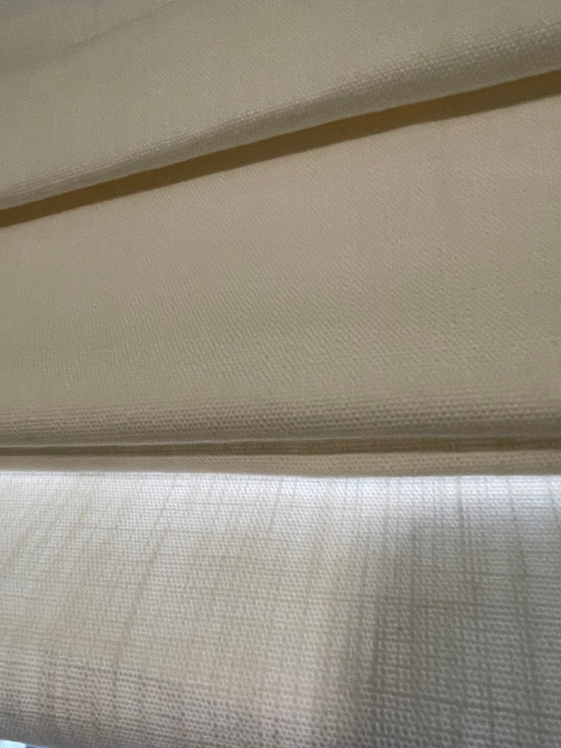 Puede incluir: Primer plano de una cortina romana de color crema, con una tela texturizada con pliegues horizontales. La cortina est&aacute; parcialmente bajada, revelando la construcci&oacute;n en capas y el sutil juego de luces y sombras en la superficie.