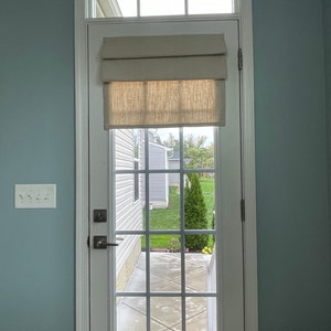 May include: A white door with a window and a beige roman blind. The door has a grid pattern and a silver handle. The window looks out onto a garden with a green lawn and a small tree.