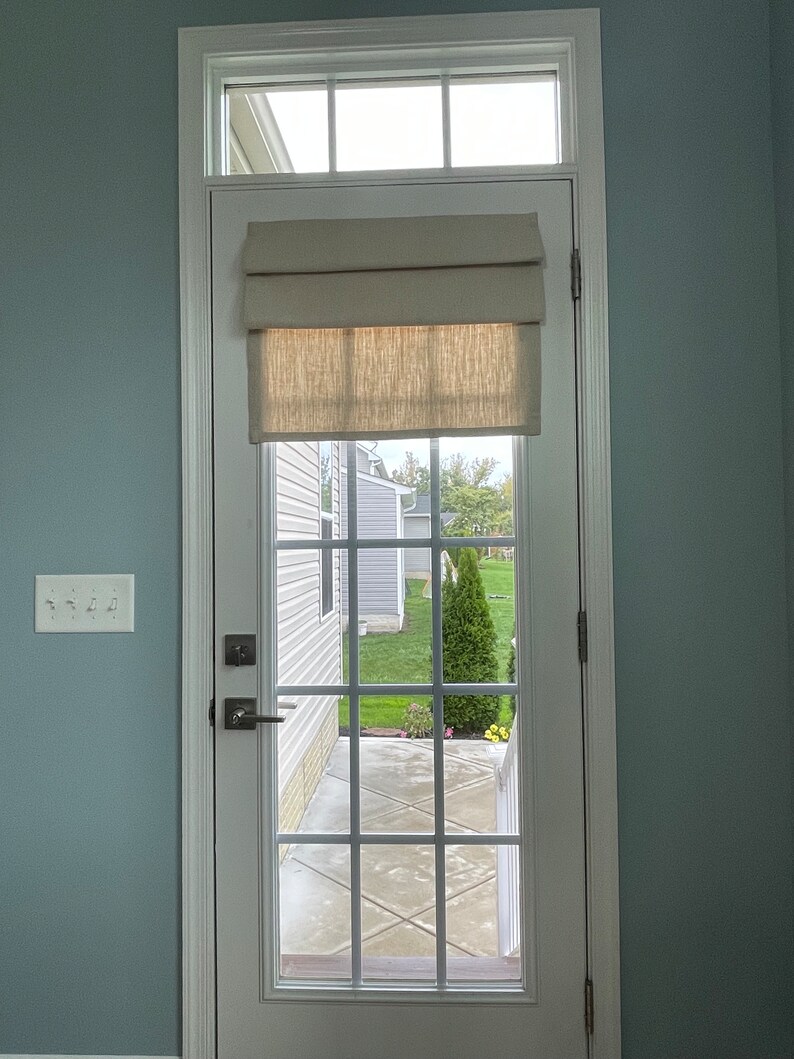 May include: A white door with a window and a beige roman blind. The door has a grid pattern and a silver handle. The window looks out onto a garden with a green lawn and a small tree.