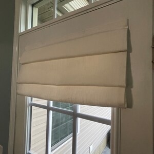 Puede incluir: Una cortina romana de color crema est&aacute; montada en una puerta blanca con una ventana. La cortina est&aacute; hecha de una tela texturizada y est&aacute; parcialmente bajada, con varios pliegues horizontales. La ventana tiene un patr&oacute;n de cuadr&iacute;cula y da a un exterior de color claro.