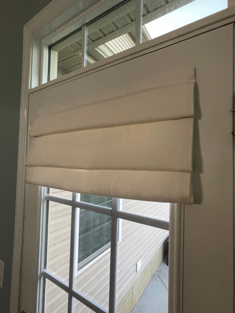 Puede incluir: Una cortina romana de color crema est&aacute; montada en una puerta blanca con una ventana. La cortina est&aacute; hecha de una tela texturizada y est&aacute; parcialmente bajada, con varios pliegues horizontales. La ventana tiene un patr&oacute;n de cuadr&iacute;cula y da a un exterior de color claro.