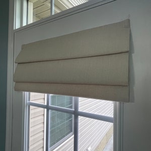 May include: A neutral-toned Roman blind is mounted on a white window frame. The blind, made of textured fabric, is partially raised, showing the window's grid and the view beyond. The window is set in a white door frame.