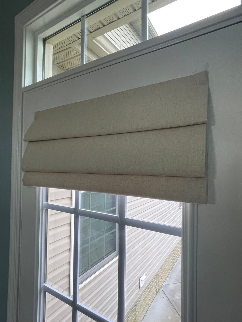 May include: A neutral-toned Roman blind is mounted on a white window frame. The blind, made of textured fabric, is partially raised, showing the window's grid and the view beyond. The window is set in a white door frame.