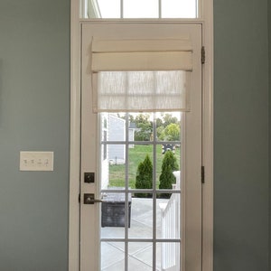 Puede incluir: Una puerta blanca con una rejilla de cristales, coronada por una peque&ntilde;a ventana y una cortina romana color crema. La puerta se abre a una vista de un patio trasero con &aacute;rboles verdes y un patio gris.