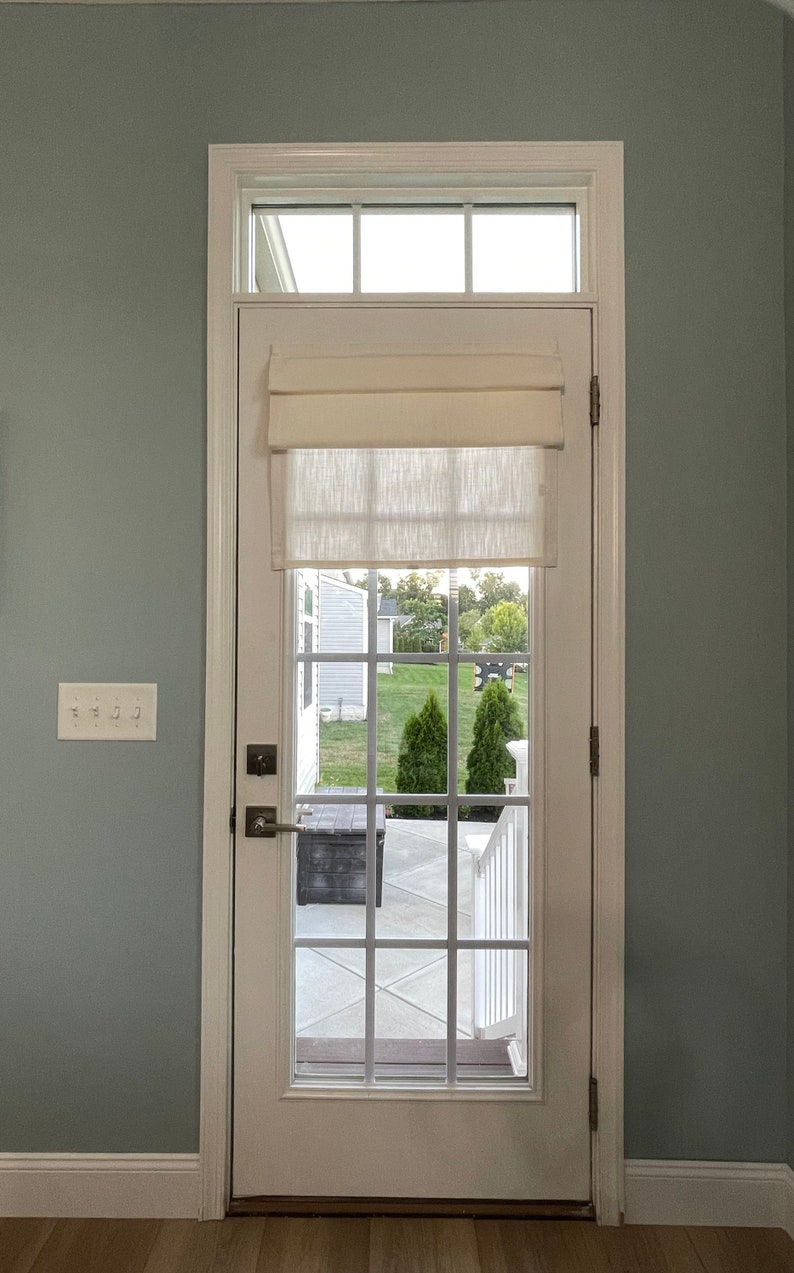 Puede incluir: Una puerta blanca con una rejilla de cristales, coronada por una peque&ntilde;a ventana y una cortina romana color crema. La puerta se abre a una vista de un patio trasero con &aacute;rboles verdes y un patio gris.