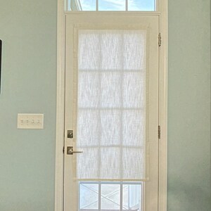Puede incluir: Una puerta blanca con una ventana rectangular y una ventana rectangular m&aacute;s peque&ntilde;a encima. Una cortina blanca, texturizada y semitransparente cubre la ventana principal. La puerta est&aacute; colocada contra una pared azul claro, con un interruptor de luz a la izquierda.