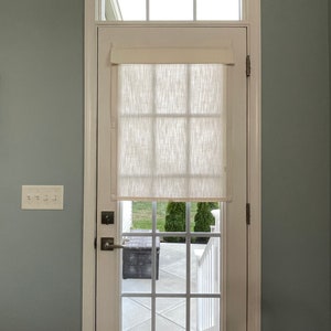 Puede incluir: Una puerta blanca con una ventana y una cortina de tela color crema. La puerta tiene un patr&oacute;n de rejilla y un tirador. La puerta est&aacute; situada en una habitaci&oacute;n con paredes azul claro y suelo de madera.