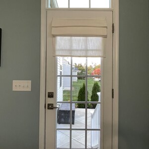 Puede incluir: Una puerta blanca con una rejilla de cristales, coronada por una ventana rectangular. Una cortina romana color crema est&aacute; parcialmente bajada. La puerta se abre a un patio con un banco oscuro y una vista de un c&eacute;sped verde y &aacute;rboles.