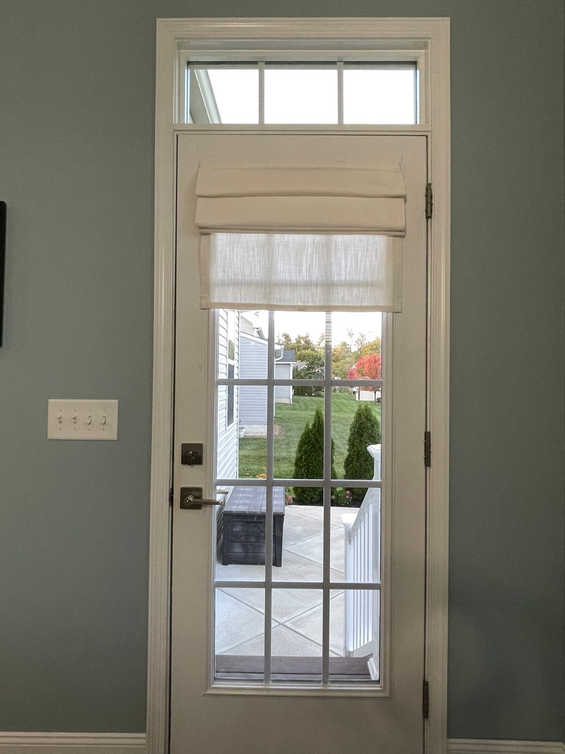 Puede incluir: Una puerta blanca con una rejilla de cristales, coronada por una ventana rectangular. Una cortina romana color crema est&aacute; parcialmente bajada. La puerta se abre a un patio con un banco oscuro y una vista de un c&eacute;sped verde y &aacute;rboles.