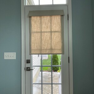 May include: A white door with a window and a beige linen shade. The door has a grid pattern and a handle. The door is set in a white frame against a light blue wall. A light switch is visible on the wall.