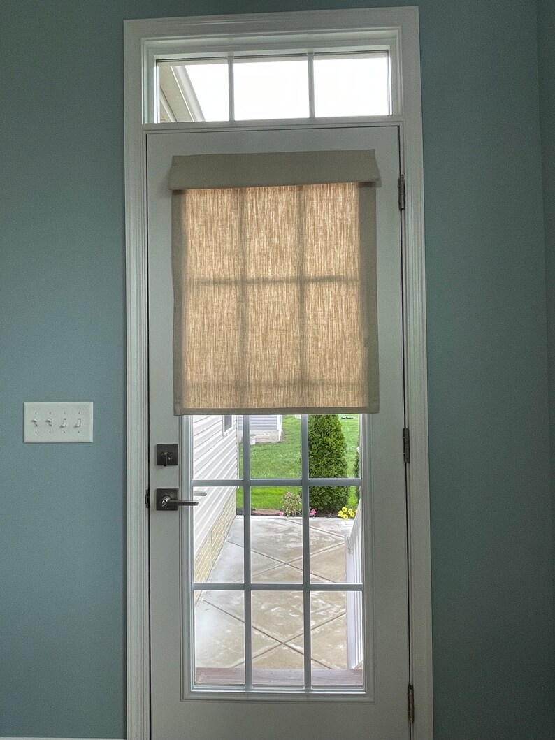 May include: A white door with a window and a beige linen shade. The door has a grid pattern and a handle. The door is set in a white frame against a light blue wall. A light switch is visible on the wall.