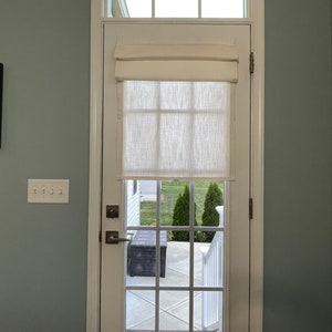 Puede incluir: Una puerta blanca con una rejilla de cristales, coronada por una peque&ntilde;a ventana. Una cortina romana color crema cubre la parte superior del cristal. La puerta se abre a un patio con un banco oscuro y vegetaci&oacute;n.