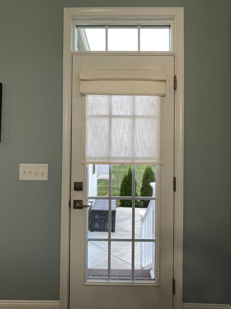 Puede incluir: Una puerta blanca con una rejilla de cristales, coronada por una peque&ntilde;a ventana. Una cortina romana color crema cubre la parte superior del cristal. La puerta se abre a un patio con un banco oscuro y vegetaci&oacute;n.