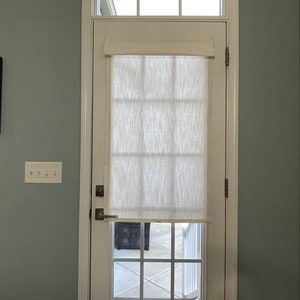 Puede incluir: Una puerta blanca con una ventana rectangular y una ventana m&aacute;s peque&ntilde;a encima. Una cortina de tela texturizada de color crema cubre la parte superior de la ventana. La puerta tiene una manija y bisagras plateadas. La pared est&aacute; pintada de azul verdoso claro.