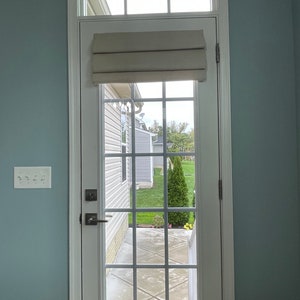 May include: A white door with a grid of glass panes, offering a view of a garden with green grass and a small tree. A beige Roman blind is mounted above the glass. The door is set in a light blue wall.